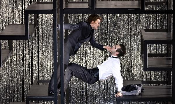 Two male actors dance, one holding a crab position as the other leans over him. They are on a metal structure.