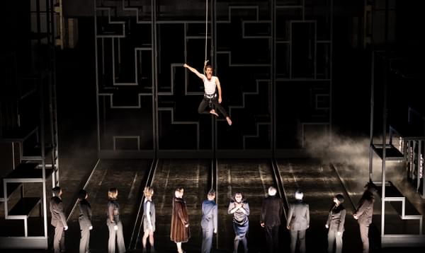 A cast of actors line up at the front of the stage. Above them hangs an actor suspended by a noose rope