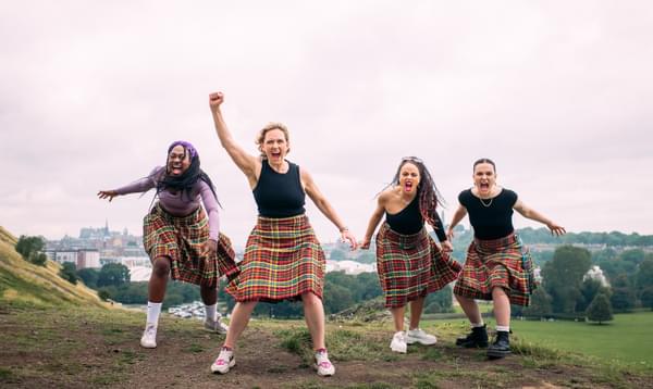 Four women lunge toward the camera screaming. They all wear black tops and kilts with red, green and yellow. Behind them are hills and green foliage.