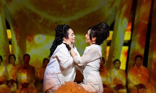 Two female actors embrace. They both wear white, and they sing passionately to each other as they hold each other.