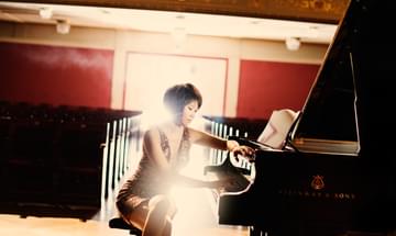 Yuja Wang sits cross-legged at a black grand piano in a grand concert hall with red walls. She is wearing a glamourous gold sequined dress and one arm rests on the piano lid while she plays piano with her right hand.
