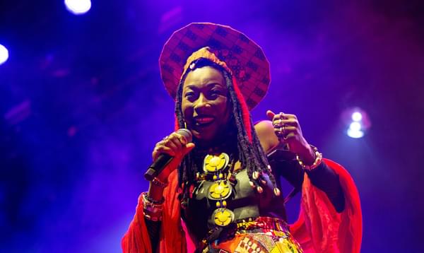 Fatoumata Diawara smiles, wearing brightly coloured clothes and hat, holding a microphone