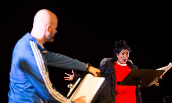 A woman speaks into a microphone with arms stretched wide, wearing a red t-shirt and winter coat. A man stands in the foreground. Both have scripts.