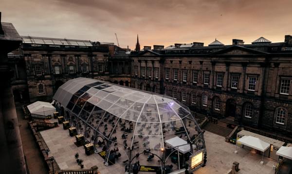 An aerial view of a large transparent tent, surrounded by the grand buildings of Edinburgh University's Old College Quad