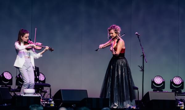 Two women play violin on stage, wearing dresses