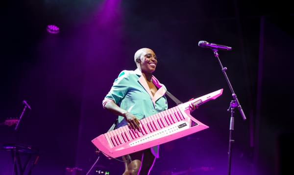 Laura Mvula stands on a dark stage wearing a pink and green blazer while smiling and playing a pink keytar.