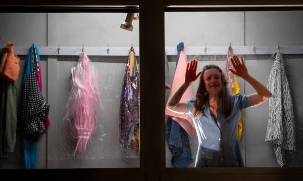 A woman press her face and hands against a window, behind her is a row of coat hangers with brightly coloured clothing hanging up