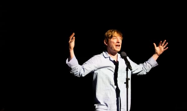 A man stands behind a microphone with his hands up in the air, wearing pyjamas