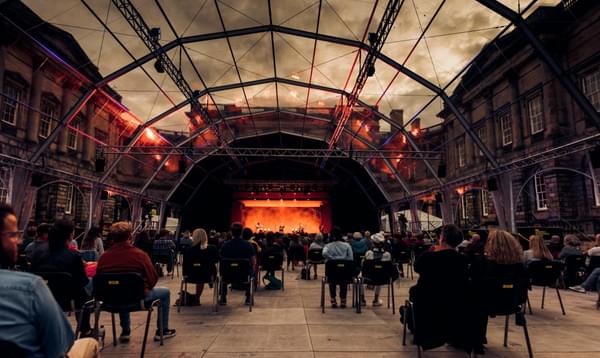 A glass-roofed tent shows cloudy skies as audience members face a colourful stage inside