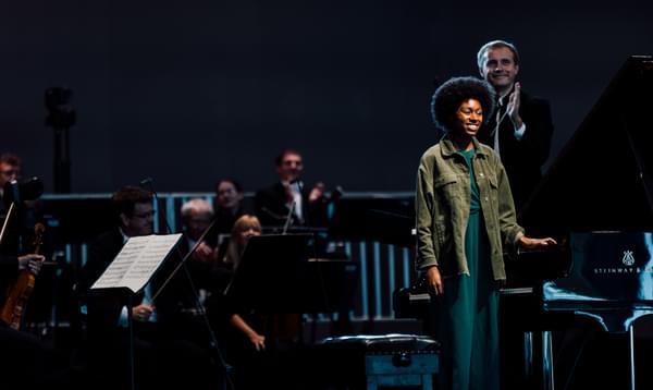 A young woman stands on stage by a piano, her afro lit up. Behind her orchestra members and conductor pose