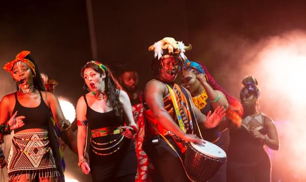 Singers and drummers wearing face paint, colourful headbands and beads perform onstage.