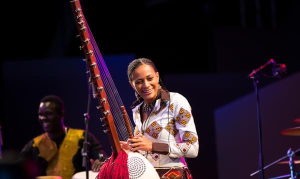 A woman stands on stage playing the kora, an African harp-like stringed instrument