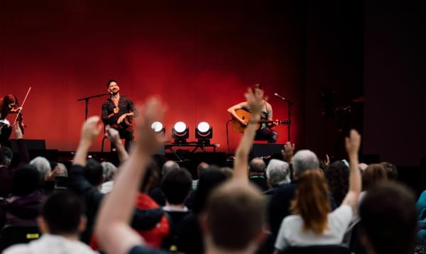 Three musicians with instruments sit on stage in the background, as audience members wave their arms in their air