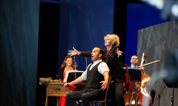 A man stands on stage in a look of shock with arms open wide, with a woman standing behind with her arms open. Behind them are a violinist and double bassist