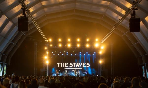 Photo taken from the back of a tent showing seated audience members in front of a stage with 'The Staves' lit up in the backdrop