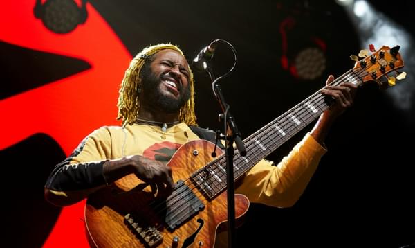 A man with bleached deadlocks plays the bass guitar with a smile or grimace
