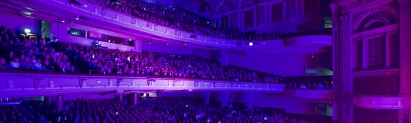 A view of the stalls, circle and upper circle of a packed theatre auditorium, bathed in a purple light.