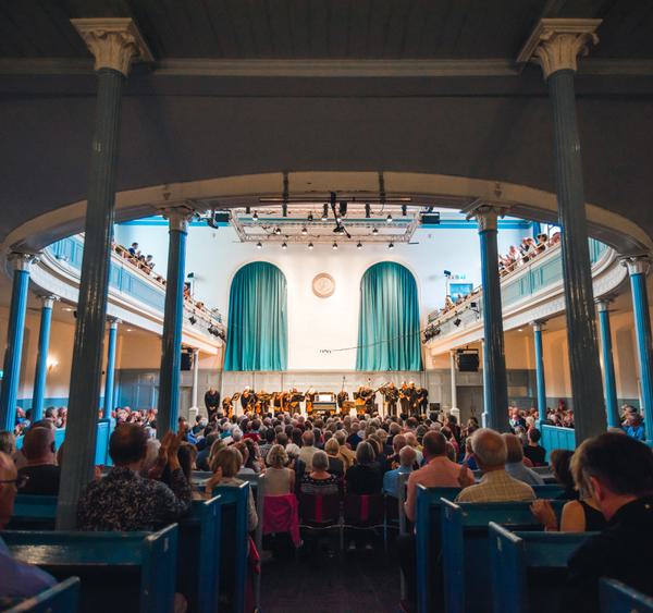 The interior of The Queen's Hall, with audience members sitting in church pews, below blue columns