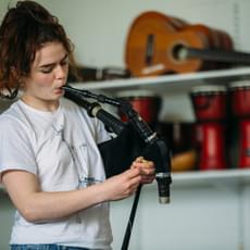 A woman in a white t-shirt with brown reddish hair piled on top of her head plays the Scottish smallpipes