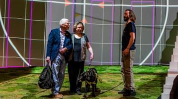 A man and woman walk arm in arm across a stage lit up with a green, grass-effect floor and geometric backdrop, talking to an actor. The woman holds the lead of a black labrador guide dog.