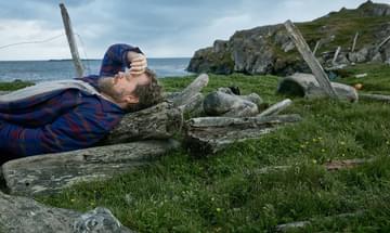 A bearded man in a grey t-shirt and blue plaid shirt pictured lying down on a clifftop, resting the back of his hand against his forehead with his eyes closed.