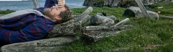 A bearded man in a grey t-shirt and blue plaid shirt pictured lying down on a clifftop, resting the back of his hand against his forehead with his eyes closed.