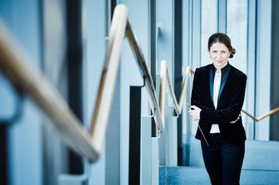 A woman stands in a modern stairwell with wooden railings and glass windows. She is smiling and wearing a black velvet suit. Her arms are crossed and she is holding a conductor's baton in her left hand.