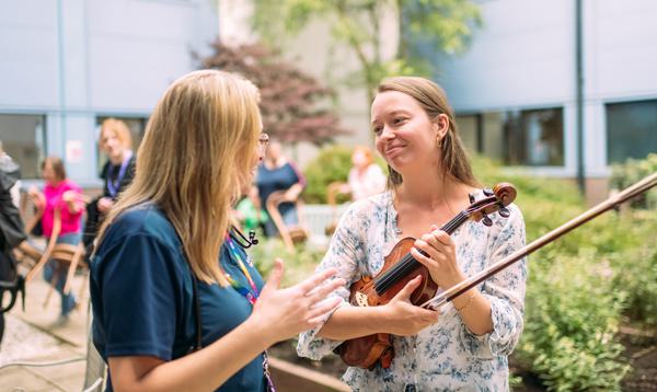 A musician holding a violin smiles as she talks to a nurse in scrubs. They are both standing outside a hospital, with green trees and plants.
