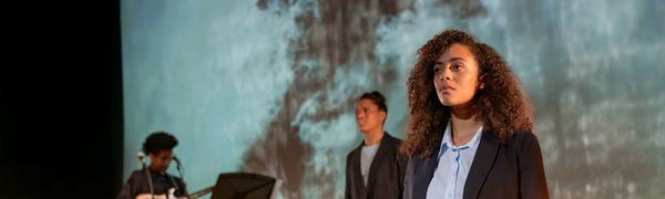 Two women stand on stage with emotional expressions, with a guitarist in the background