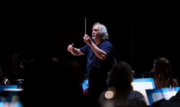 A man with grey curly hair stands in a blue shirt against a black background, holding a conducting baton
