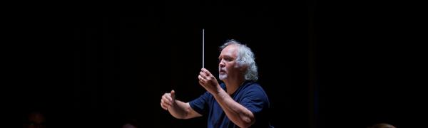 A man with grey curly hair stands in a blue shirt against a black background, holding a conducting baton