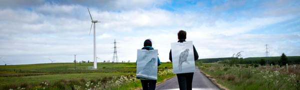 A man and a woman walk down a road between two grassy fields, carrying pictures of owls on their backs