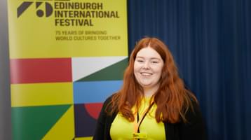 A woman with red hair in a yellow t-shirt smiles in front of an International Festival banner