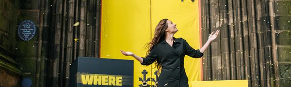 A woman standing in front of grand yellow doors holds her hand out and looks up to the side smiling as yellow confetti swirls in the air around her.