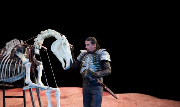 A man in skeletal armour stands talking to a life-size skeleton of a horse