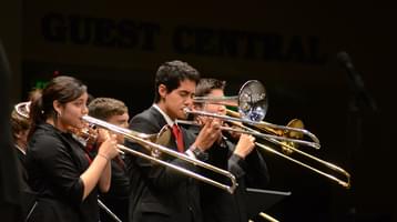 Young trombone players dressed in black shirts perform against a black backdrop, with other brass instruments visible in the background.