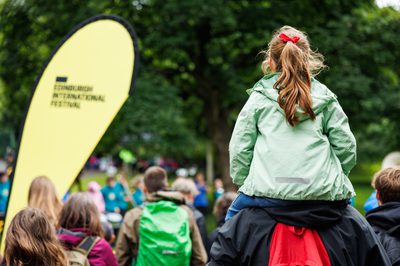 A child wearing a red bow sits on the shoulders of an adult, while standing in a crowd.