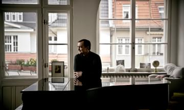 A man stands in front of large windows leaning against the top of a grand piano