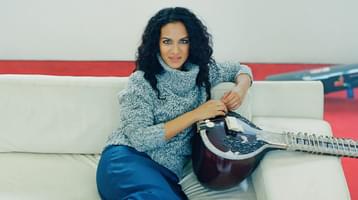 The artist pictured wearing a cosy knit jumper sitting on a white sofa, leaning one arm against the back of it with a sitar on her lap.