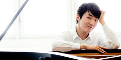 The pianist shown wearing a white shirt, looking at the viewer over the open lid of a grand piano.
