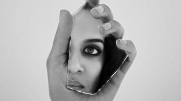 A black-and-white photo of a hand holding a fragment of shattered mirror, with part of the artist's face visible in the reflection.