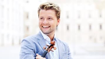A broadly smiling man with dark blonde hair, wearing a blue striped shirt and pale blue jacket, holding a violin against his chest.