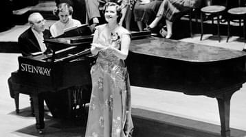 A singer wearing a long gown stands onstage in front of a grand piano, looking upwards and holding a small notebook in her hands.