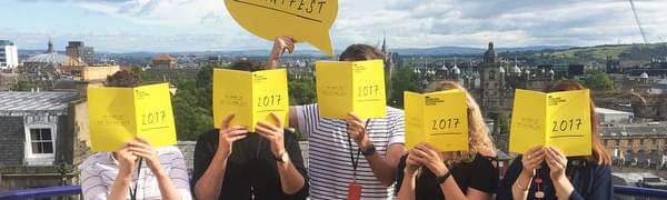 Five people standing on a balcony with the Edinburgh skyline visible behind them, each covering their face with an open copy of the 2017 Edinburgh International Festival brochure.