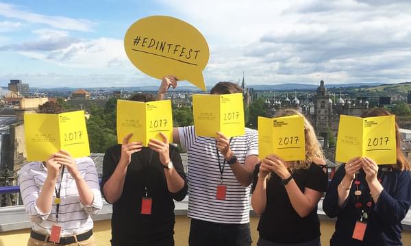 Five people standing on a balcony with the Edinburgh skyline visible behind them, each covering their face with an open copy of the 2017 Edinburgh International Festival brochure.