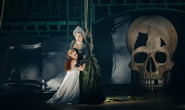 A woman in a white gown kneels clinging to the skirts of another elaborately dressed woman, with a large skull looming out of the darkness behind them