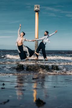 Standing in the sea water before a tall marker, a dancer stands side-on in an arabesque, his arm reaching above his head. He is supported by another dancer, who holds his hand, counter-balancing him as he reaches behind him.