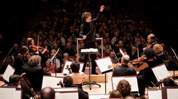 A view over the musicians' heads of a conductor onstage in mid-performance, raising his left hand. The seated audience is visible in the background.