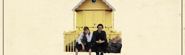 A young man and woman sit looking bored on the steps in front of a small yellow house