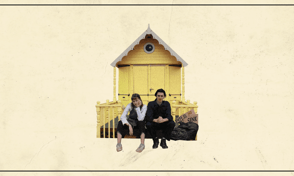 A young man and woman sit looking bored on the steps in front of a small yellow house